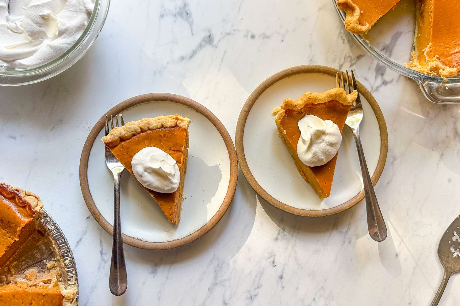 Two slices of pumpkin pie served on plates with a dollop of whipped cream each accompanied by forks on a marble countertop