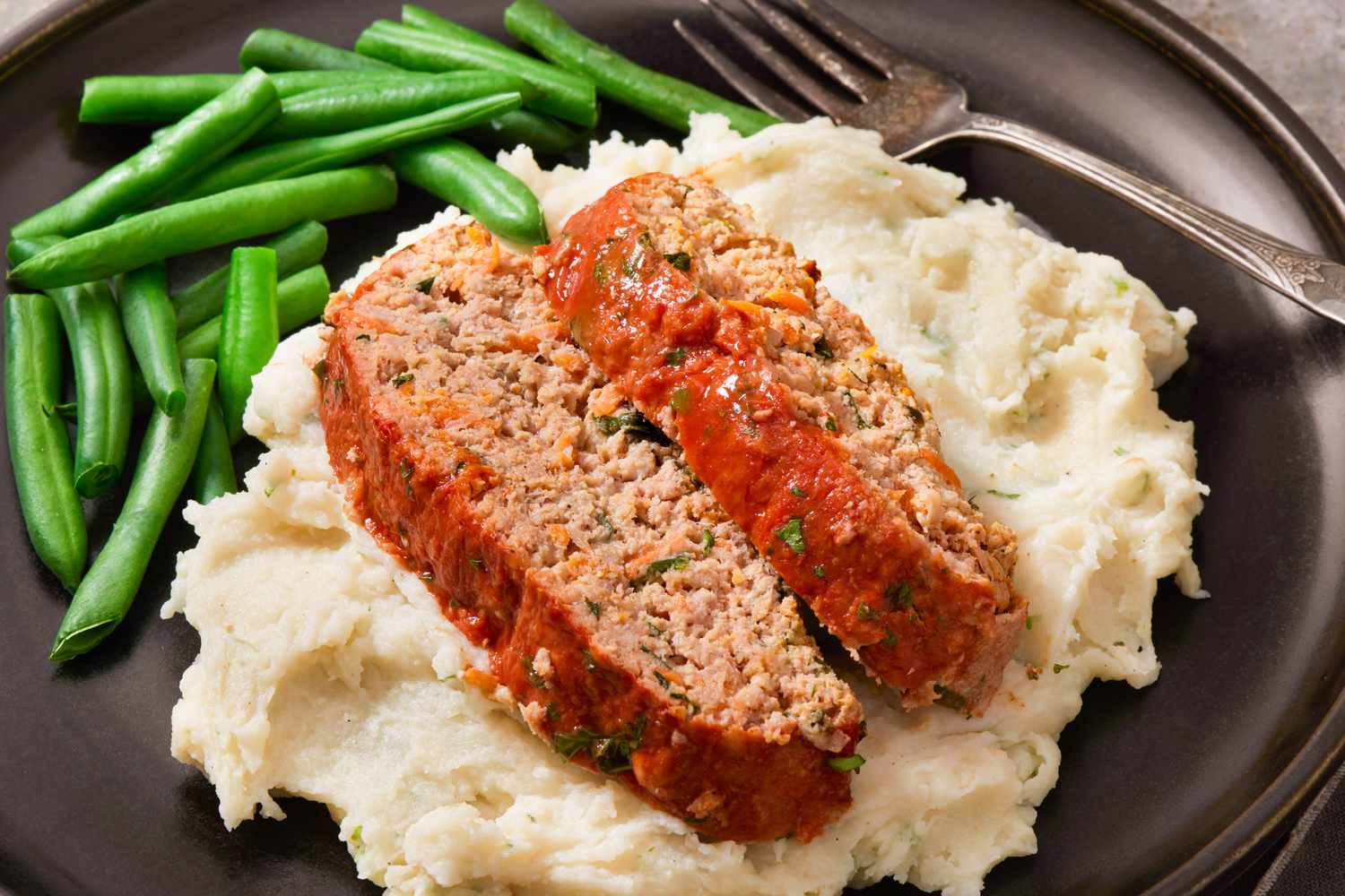 A plate with two slices of meatloaf on top of mashed potatoes and a side of green beans