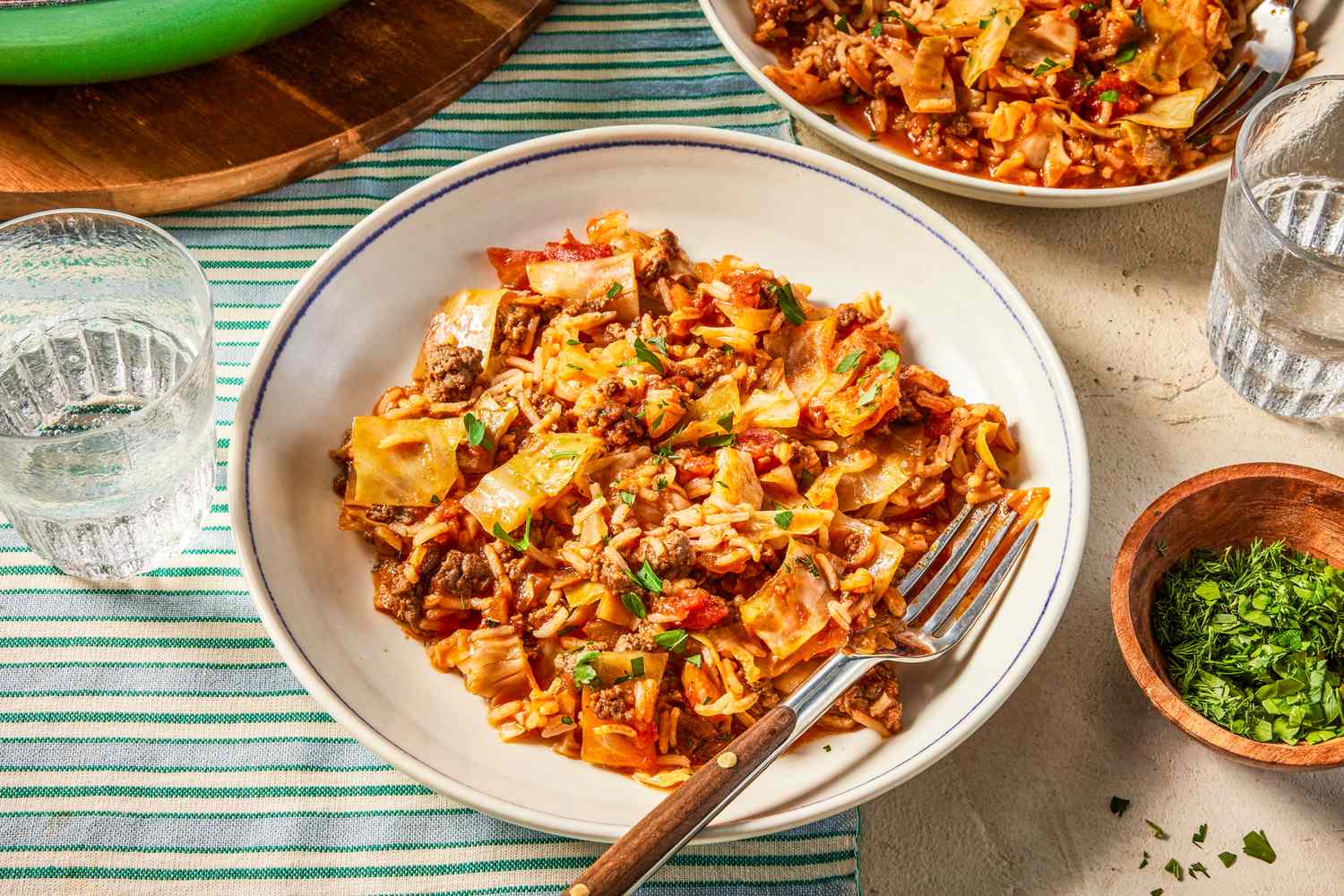 unstuffed cabbage rolls served on a plate at a table setting with another serving on a plate, a small bowl of minced herbs, glasses of water, and more of recipe in a pan