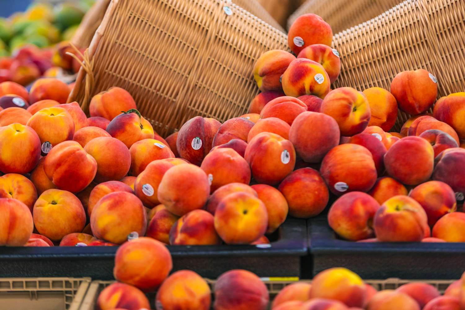 A pile of peaches in a grocery store, baskets of other fruits in the background. Some of the peaches have visible stickers