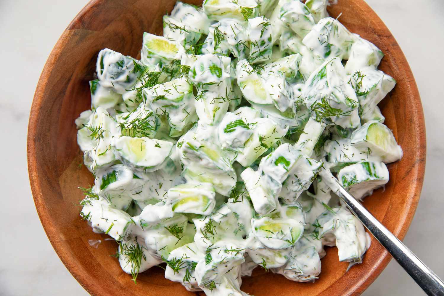 Cucumber salad in a wooden bowl with a creamy dressing and dill, served with a spoon
