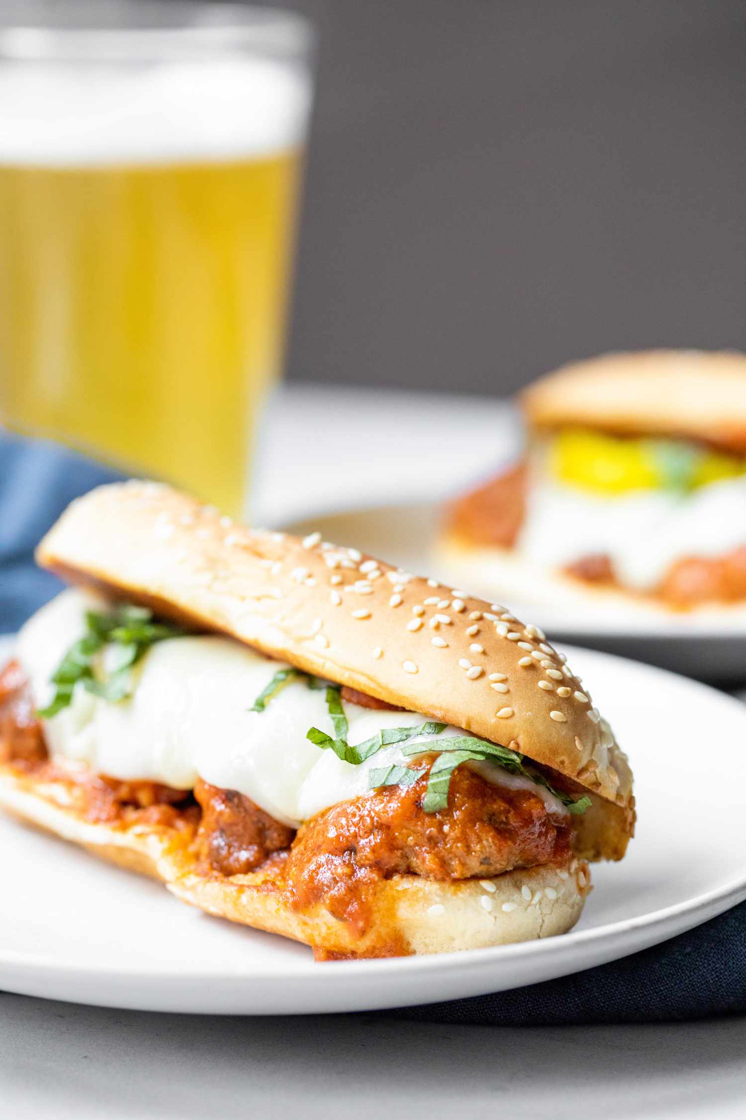 A Plate of Meatball Sub Topped with Banana Peppers and Basil, and in the Background, a Plate With Another Sub and a Glass of Beer