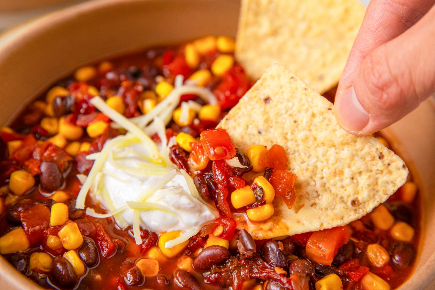A bowl of chili with corn beans tomatoes sour cream and shredded cheese with a hand dipping a tortilla chip into it