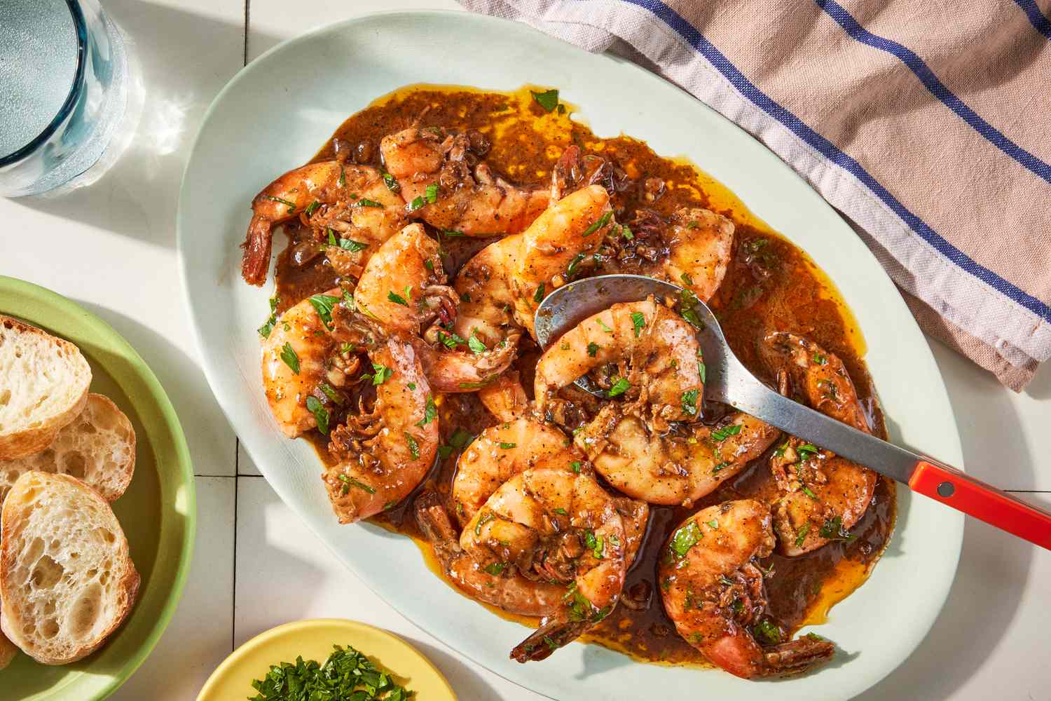 New Orleans BBQ Shrimp in a platter at a table setting with a bowl of sliced baguettes, bowl of herbs, a glass of water, and table napkin