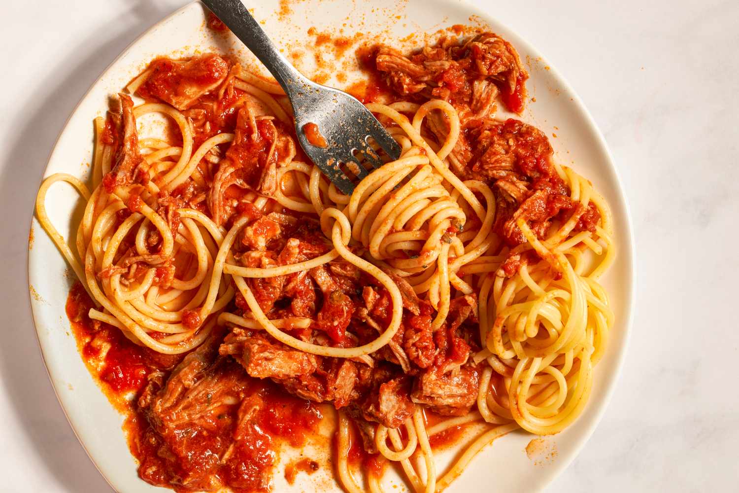 Close-up of a plate of spaghetti with tomato sauce and shredded meat. A fork is coming in and has a twirl of pasta around it.