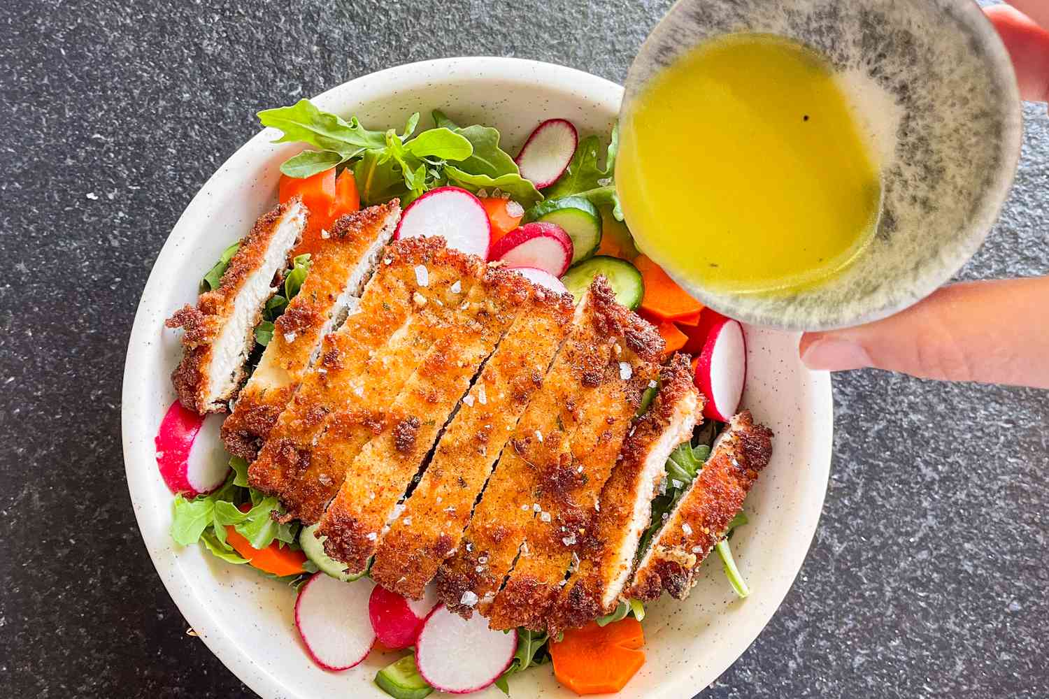 Overhead view of a white bowl of sliced fried chicken on a bed of greens, carrots, radishes and cucumbers with a bowl of dressing being poured over the salad all on a gray background