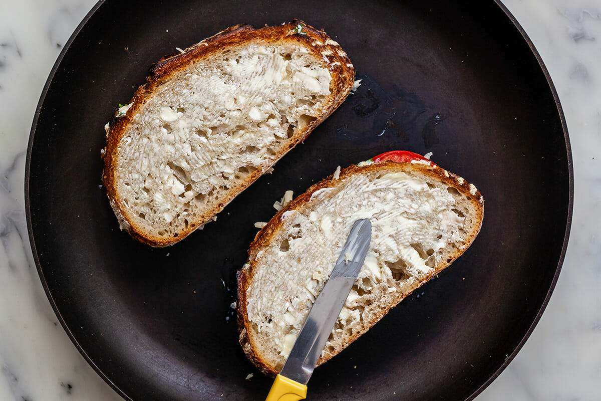 Two tuna melt sandwiches in a cast iron skillet. The tops are being spread with butter.