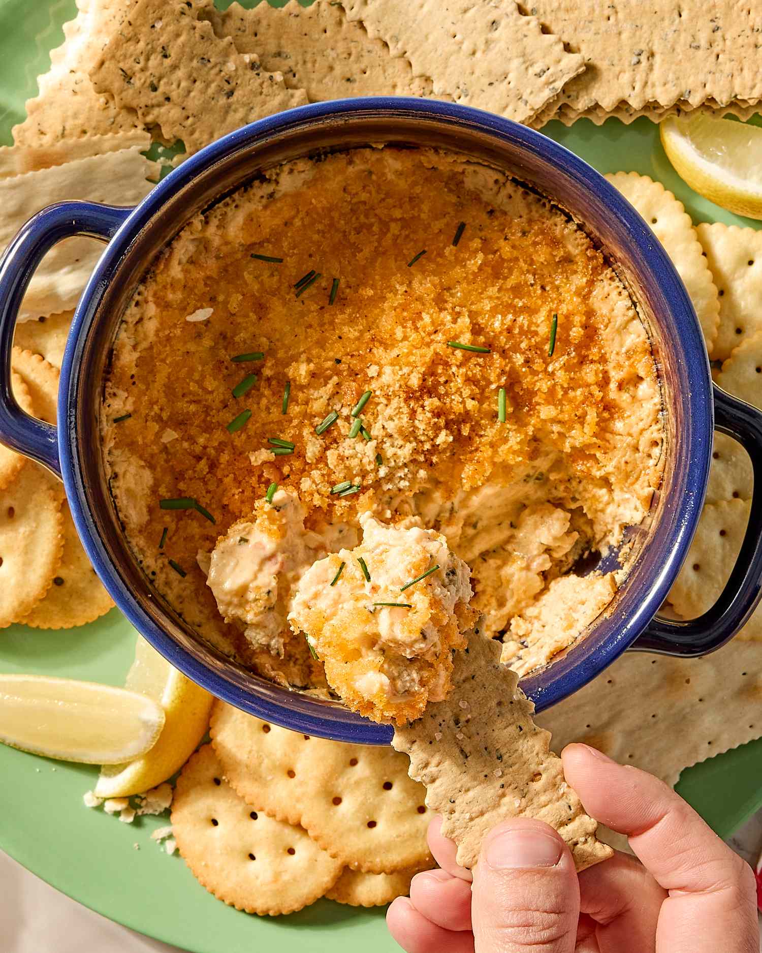 A dish of lobster dip with crackers and a hand dipping a piece of cracker