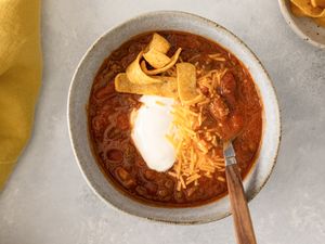 Bowl of Grandma Patty’s Chili with sour cream shredded cheese and Fritos alongside a bowl of Fritos and a yellow napkin