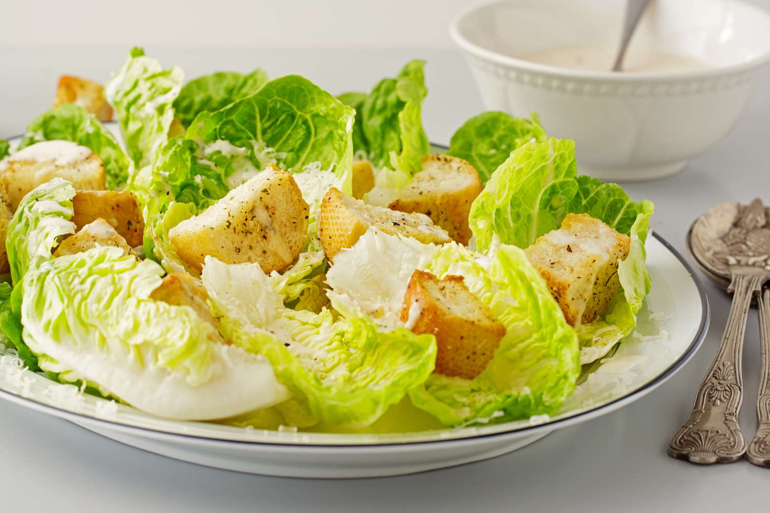 A Caesar salad with romaine lettuce, croutons, and grated cheese on a white plate, with a bowl and utensils in the background