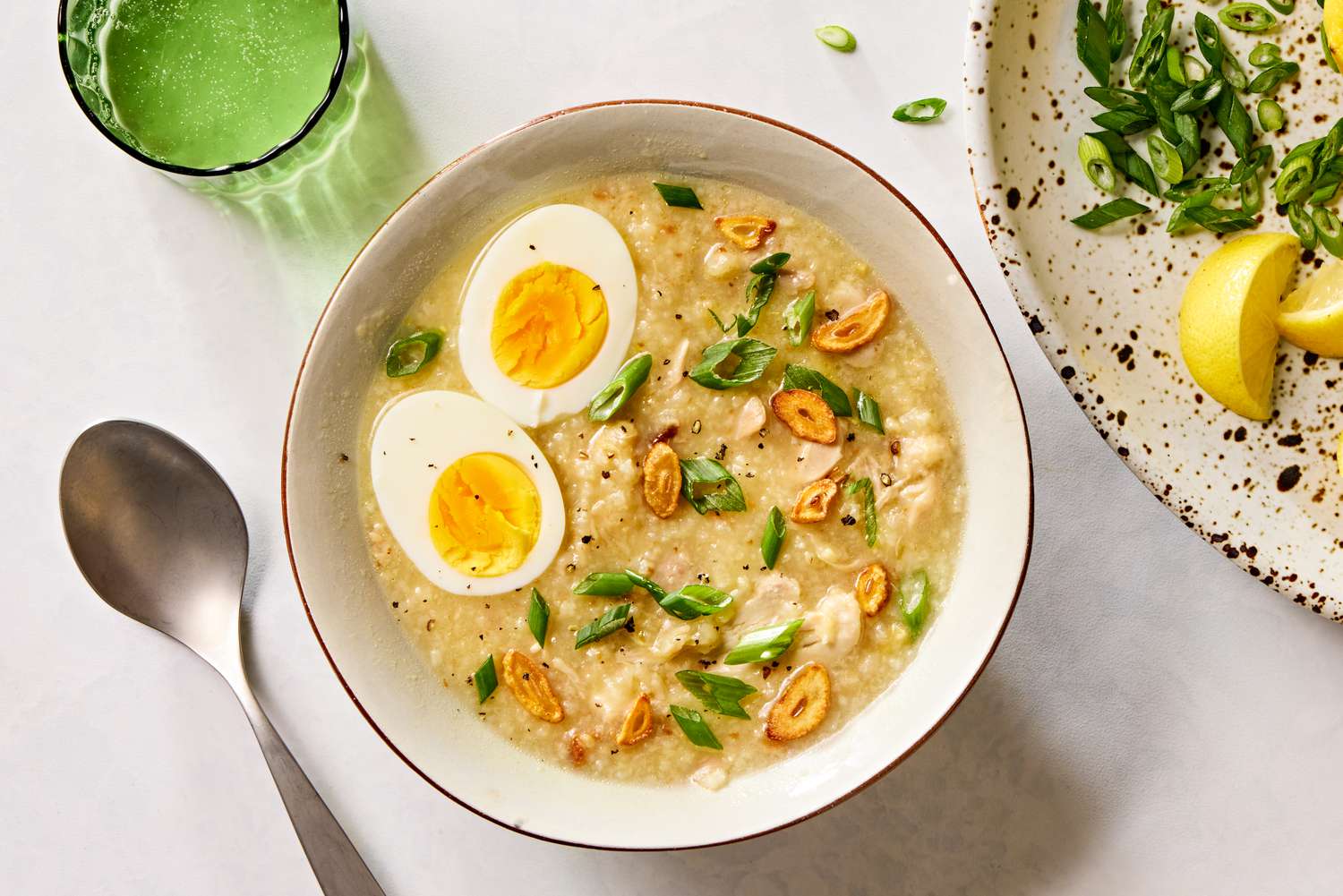 Overhead view of a bowl of Arroz Caldo with Chicken recipe next to a spoon, green glass and plate of lemon slices and scallions 