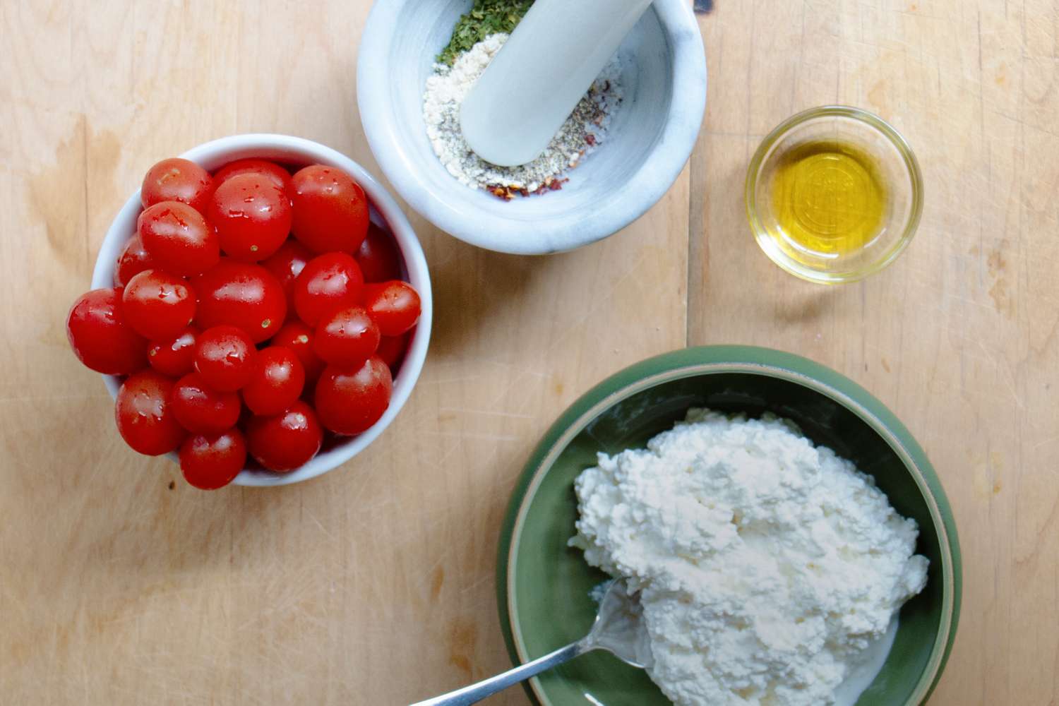 Ingredients on a wooden surface include a bowl of cherry tomatoes a bowl of cottage cheese a mortar with spices and a small glass of olive oil