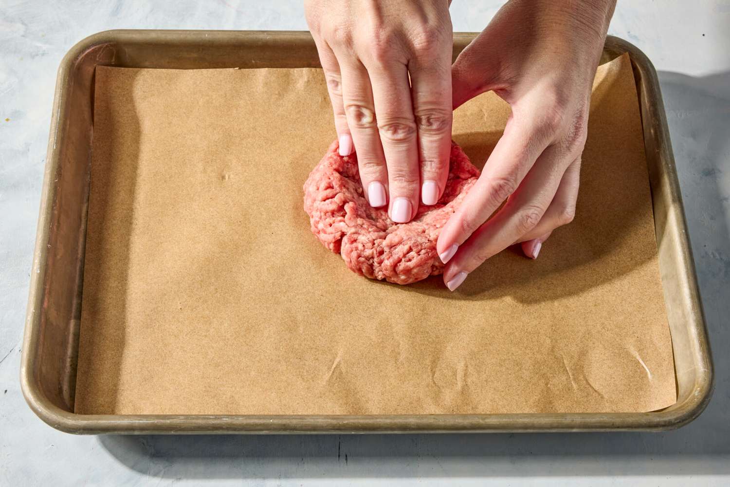 Hands shaping ground meat into a burger patty on a parchment-lined baking tray