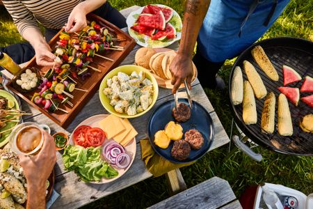 Outdoor meal with skewers, burgers, and grilled items on a picnic table near a grill