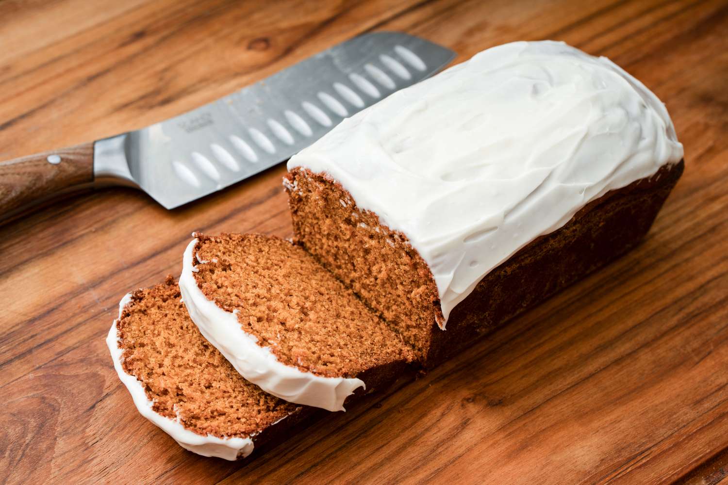 A frosted loaf cake with a few slices cut on a wooden surface next to a knife