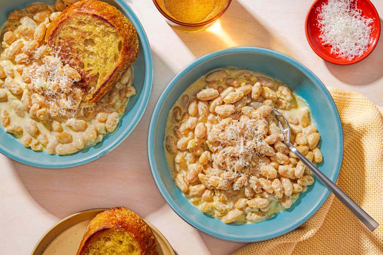 Overhead view of two teal colored bowls of Alfredo Beans, one with a spoon and one with a piece of bread, next to a plate of bread, drinking glass and small red bowl of shredded Parmesan