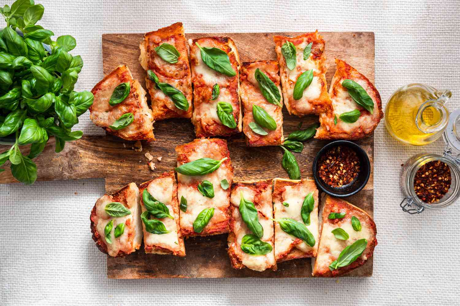 Sliced French Bread Pizza Topped With Fresh Basil (Unsliced) and a Small Bowl of Crushed Peppers on a Wooden Platter and on the Counter Next to It, an Olive Oil Dispenser, a Jar With More Crushed Red Peppers, and a Bushel of Fresh Basil