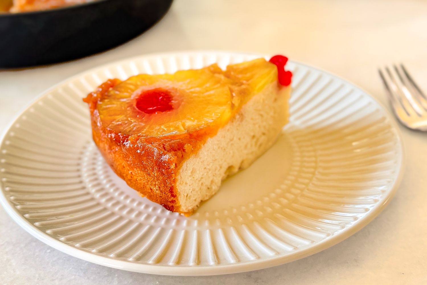 A slice of pineapple upsidedown cake on a plate garnished with a cherry and pineapple ring a fork in the background
