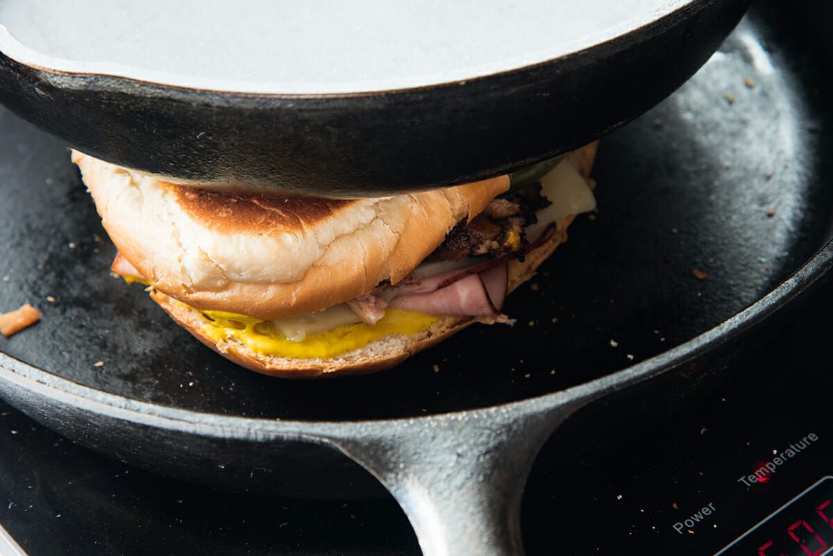 Cast iron skillet on a stove with a traditional cuban sandwich pressed between another cast iron skillet. Toasted bread, mustard, pork and cheese showing.