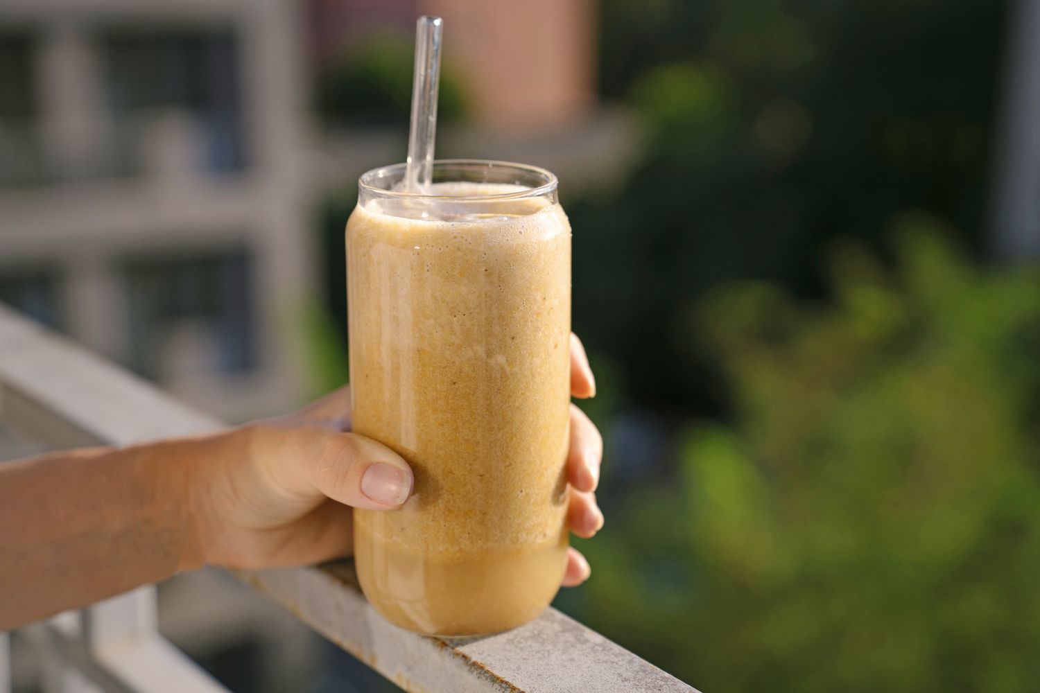 A hand holding a glass filled with a blended smoothie and a straw outdoors on a sunny day