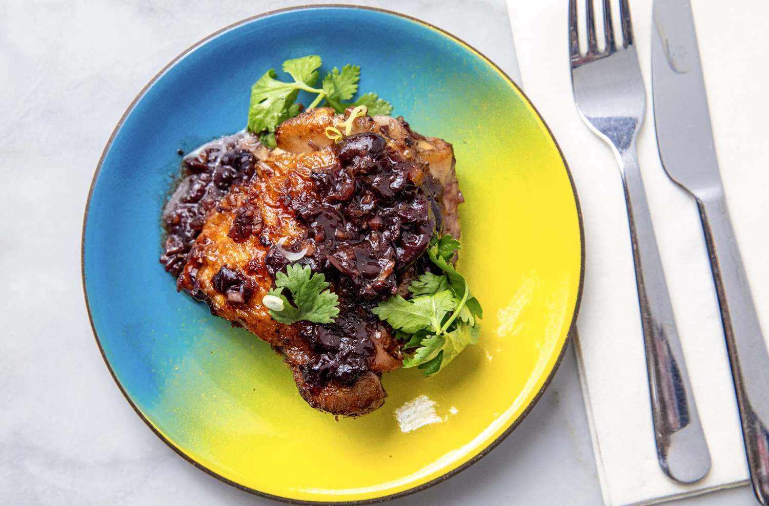 A plate with sticky chicken garnished with fresh herbs and a dark sauce knife and fork beside it on a napkin