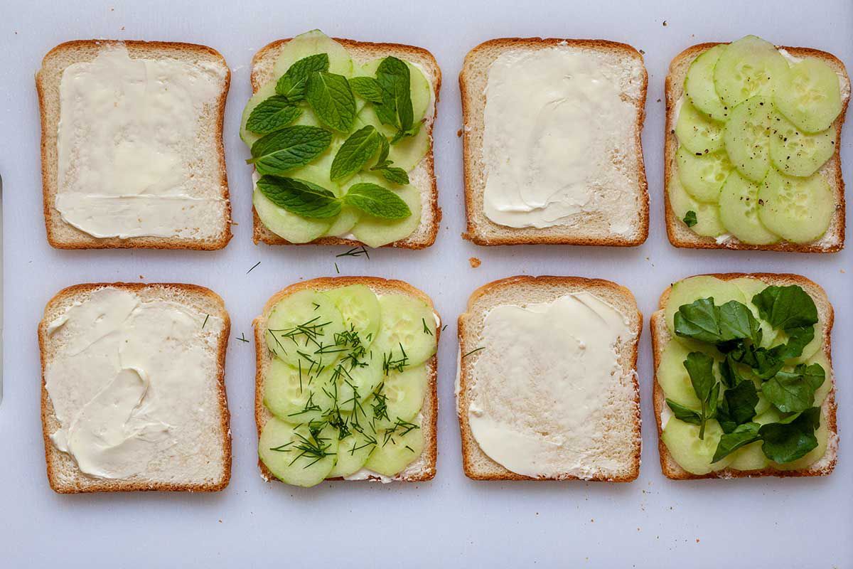Eight slices of bread in two rows of four. Cream cheese is spread on every other slice and cucumbers with herbs are on the other slices to make cucumber cream cheese sandwiches.
