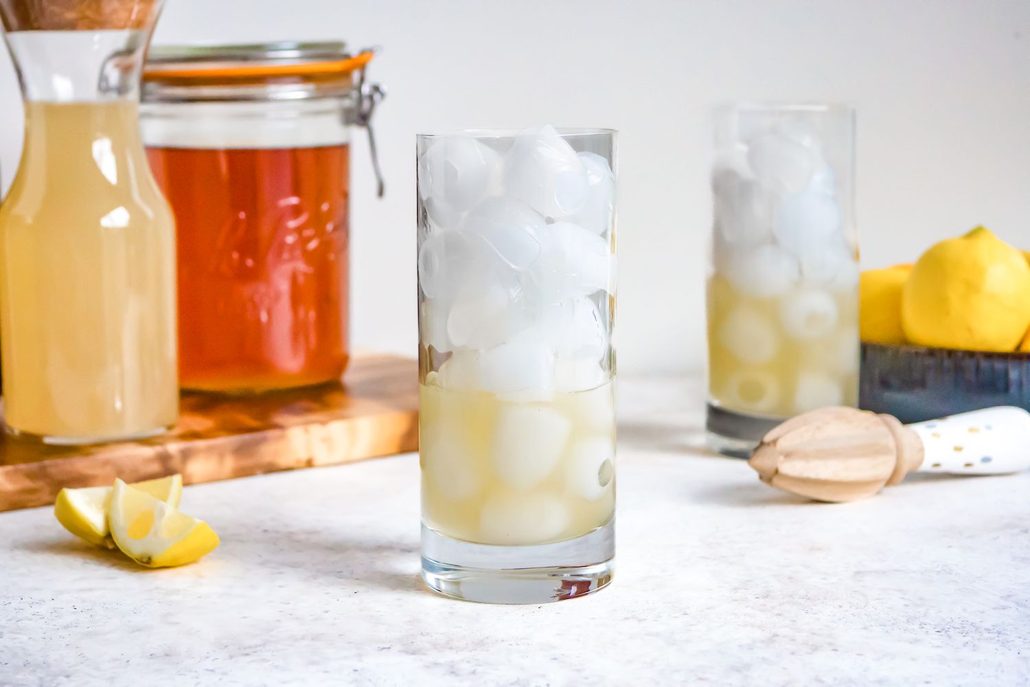 Glass Filled With Ice and Some Lemonade, and in the Background, Another Glass With Ice and Lemonade, a Bowl of Lemons, and a Citrus Reamer. On the Left, Two Jars (One With Lemon Juice and Another With Black Tea) on a Wooden Board and Two Lemon Wedges for Arnold Palmer Recipe