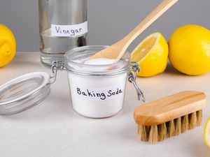 A glass jar of baking soda and a bottle of vinegar on a counter, with a cleaning brush and lemons scattered around