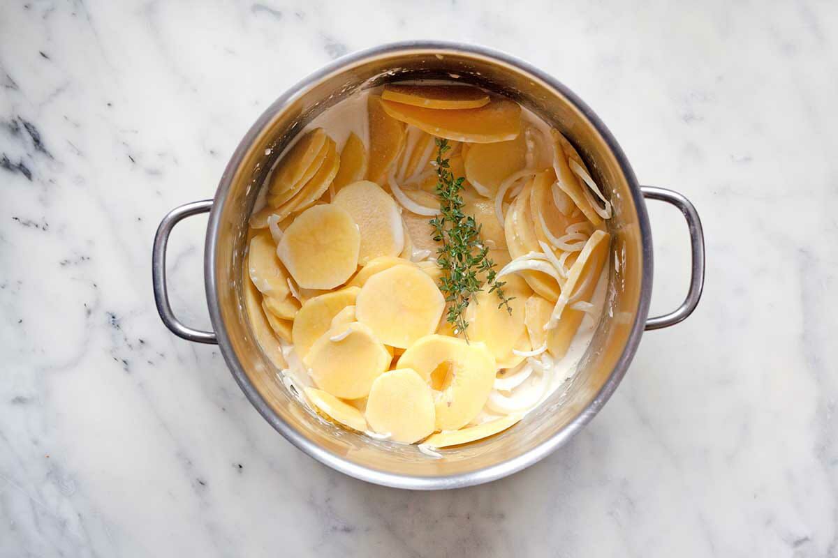 Au Gratin Potatoes are being made in a pot. Fresh thyme sits on top of thinly sliced potatoes nestled in a cream sauce.