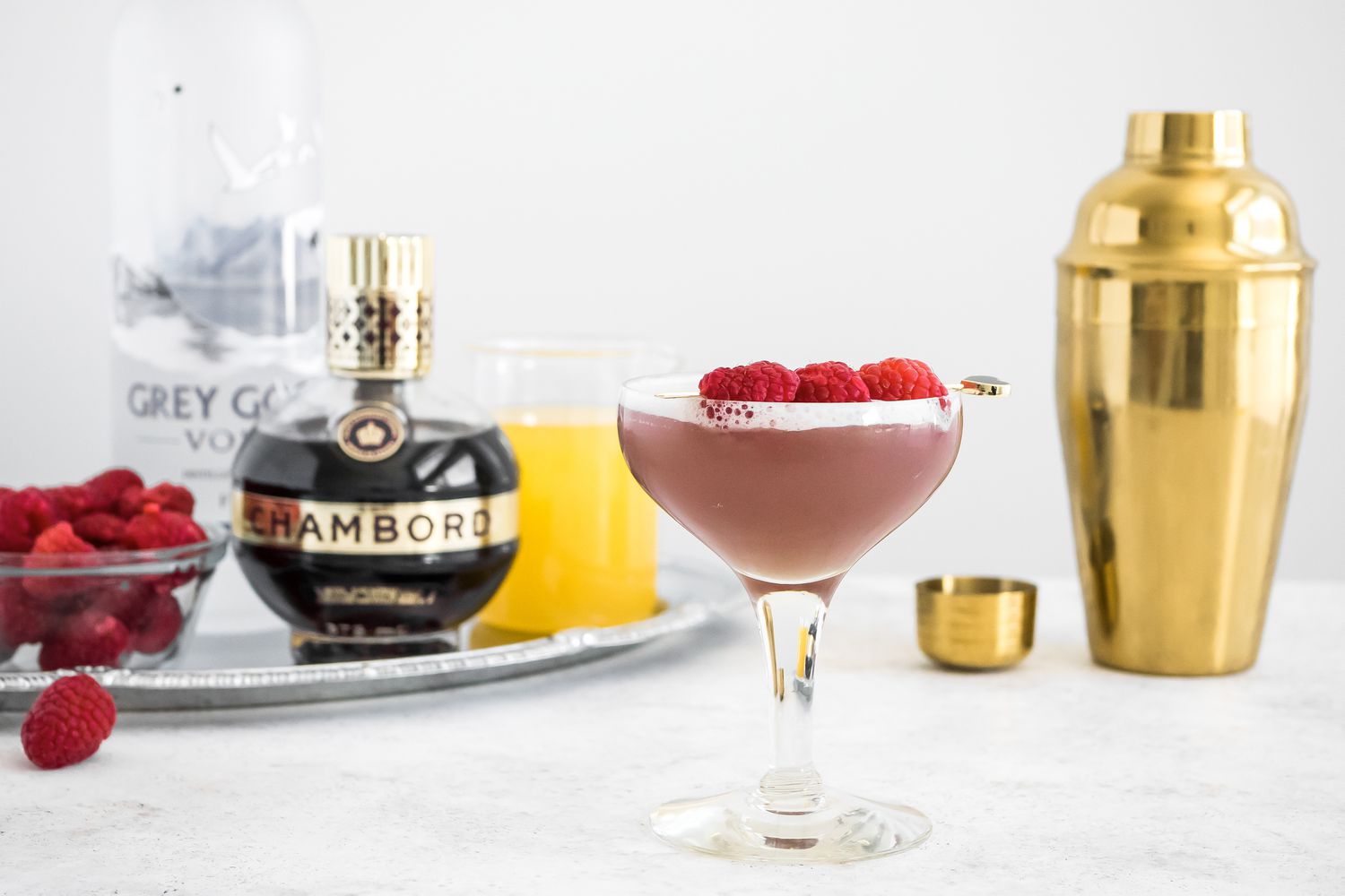 French Martini in a Glass Garnished with Raspberries and Surrounded by Shaker and a Tray with a Bowl of Raspberries, Vodka, Chambord, and Pineapple Juice 