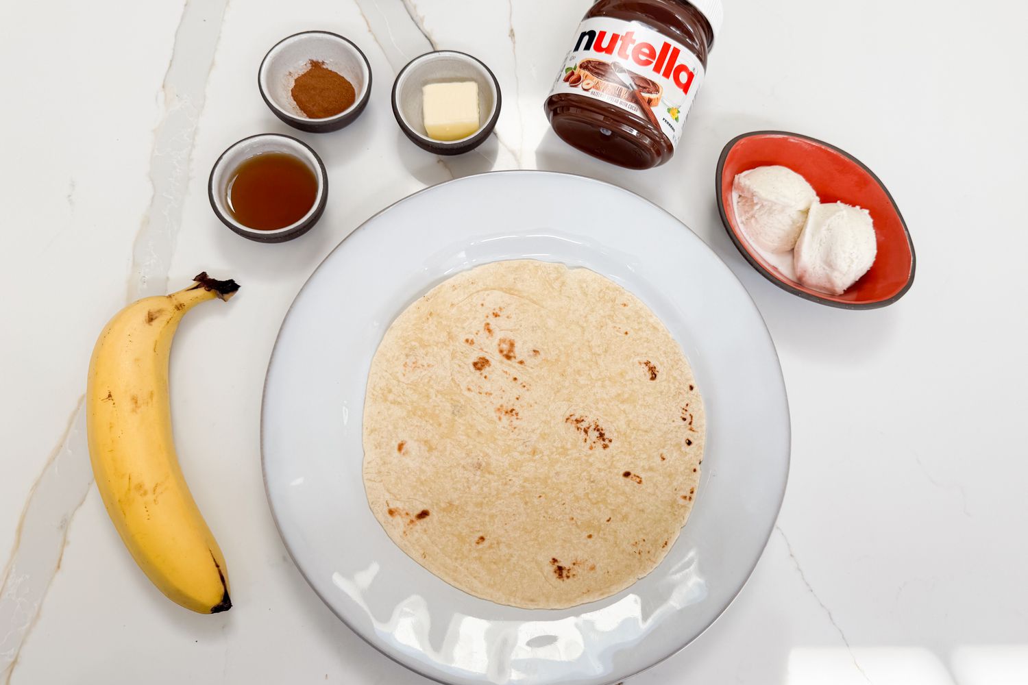Ingredients for making tortilla dessert with Nutella, banana, cinnamon, ice cream, and butter on a white surface