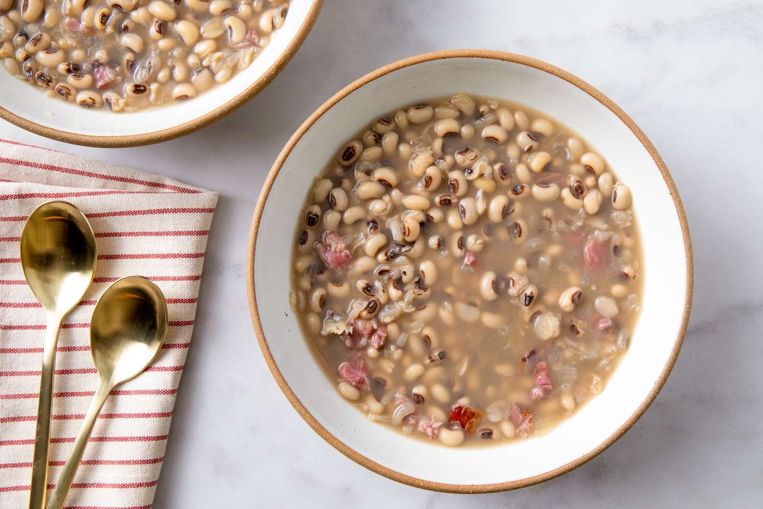Two bowls of blackeyed peas soup served with gold spoons on a striped cloth napkin