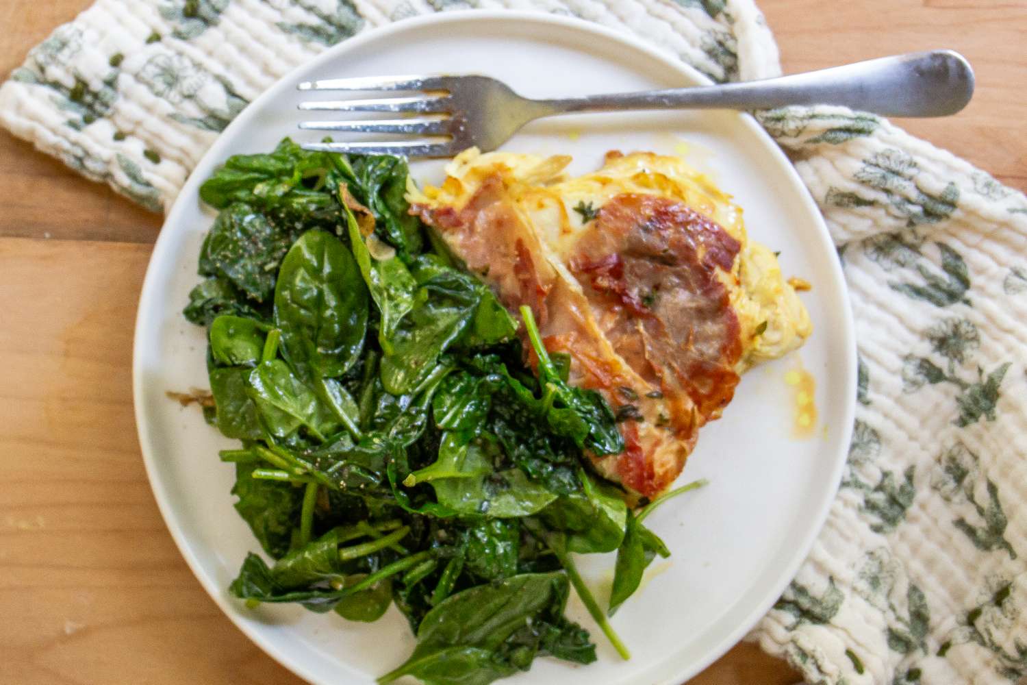 A plate with sauted greens and a serving of a layered dish featuring thinly sliced meat served with a fork on the side