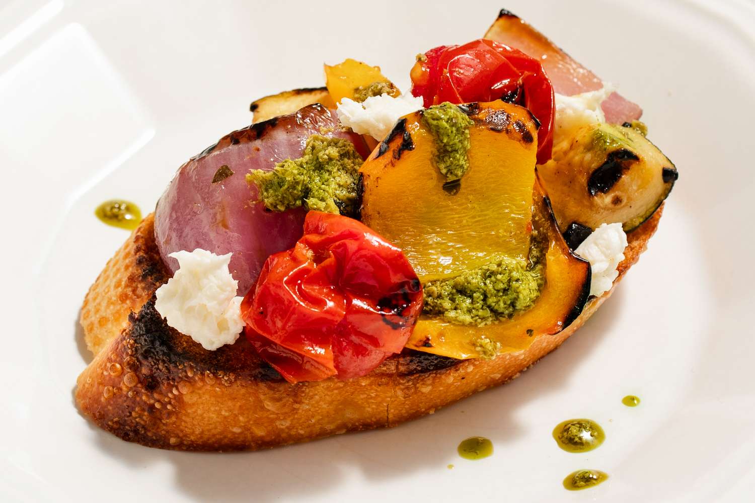 Grilled bread topped with roasted vegetables, dollops of cheese, and a drizzle of green sauce