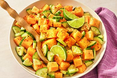 Bowl of cantaloupe pieces sliced cucumbers lime wedges and Tajn seasoning wooden spoon purple napkin nearby