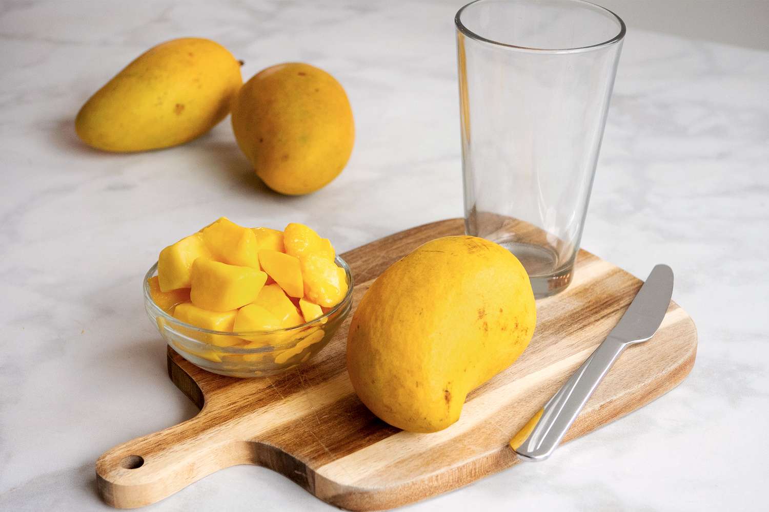 Sliced mango pieces in a bowl on a cutting board with a whole mango a glass and a knife nearby