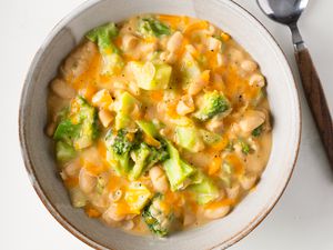Overhead view of a stoneware bowl of Broccoli Cheddar Creamy Beans next to a spoon and all on a white background