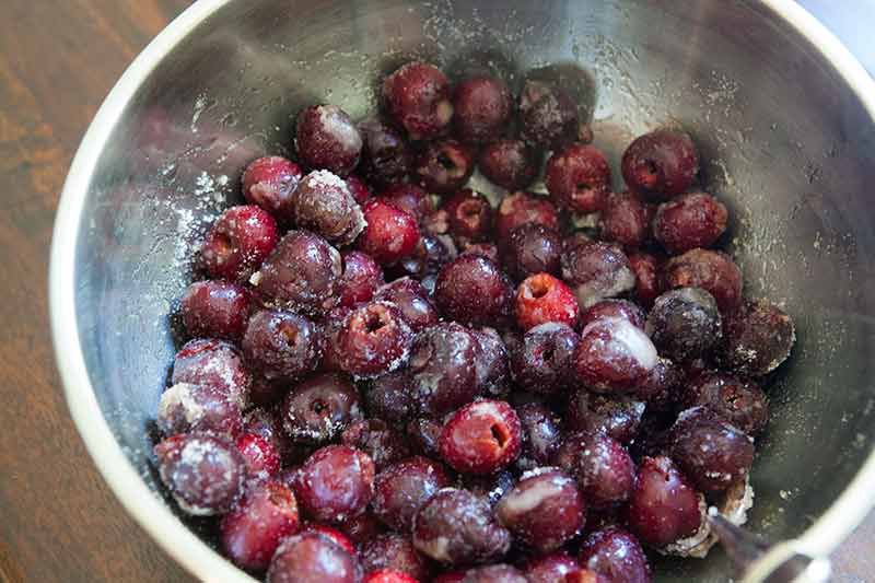 A metal bowl with fresh cherries coated in sugar show how to make cherry pie.
