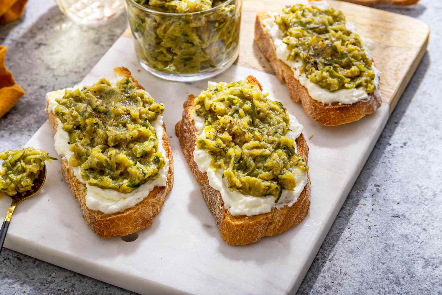 Slices of Toast Topped With Ricotta and Zucchini Butter on a Marble Slab Next to a Bowl With More Zucchini Butter and a Spoon, and in the Surroundings, a Mustard Colored Kitchen Towel and a Glass