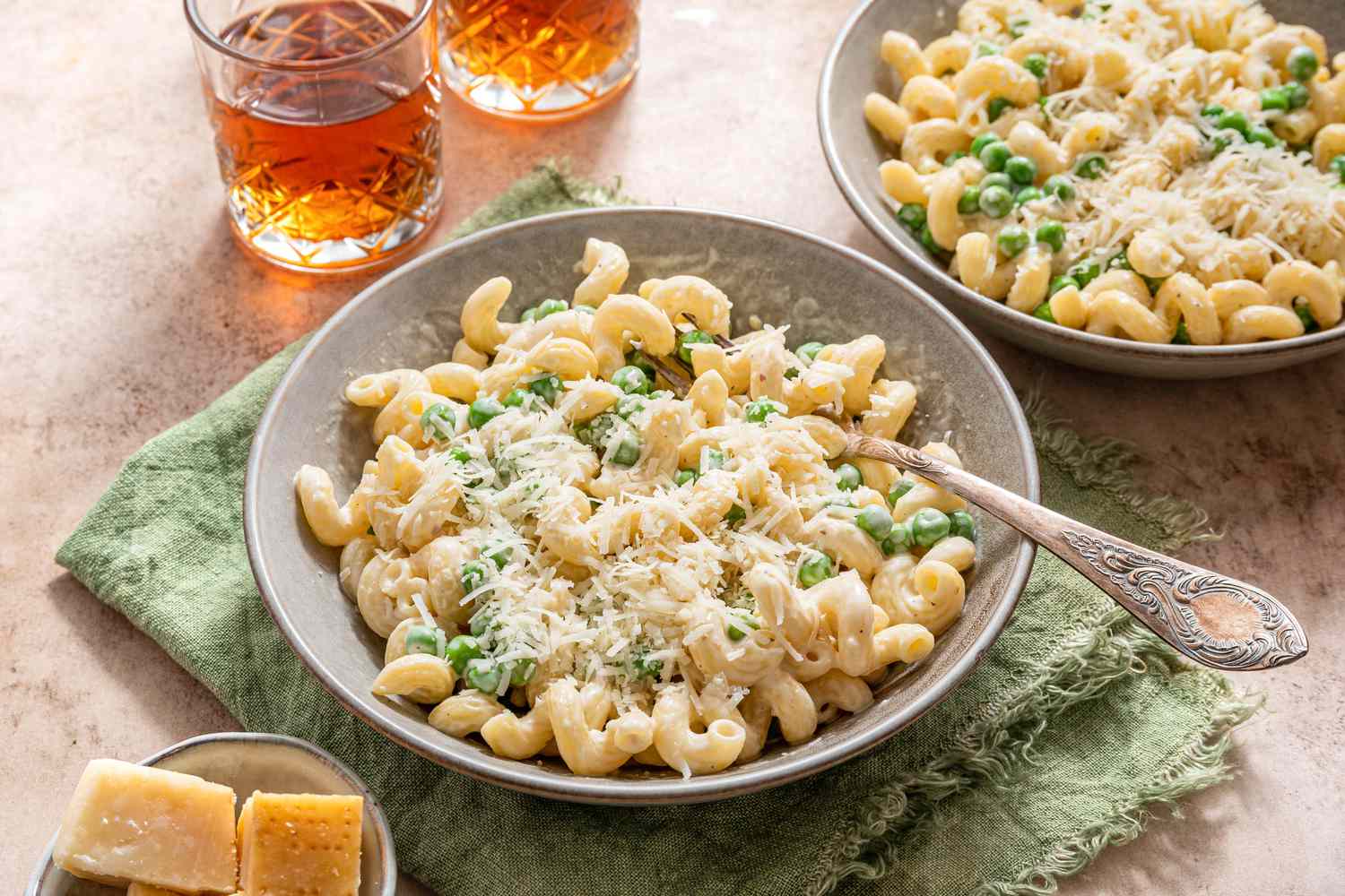 Bowl of Creamy Cavatappi Topped With Shredded Parmesan With a Fork and Sitting on an Olive Kitchen Towel. In the Surroundings, Another Bowl With a Serving, a Small Bowl With Blocks of Parmesan, and a Two Glasses.