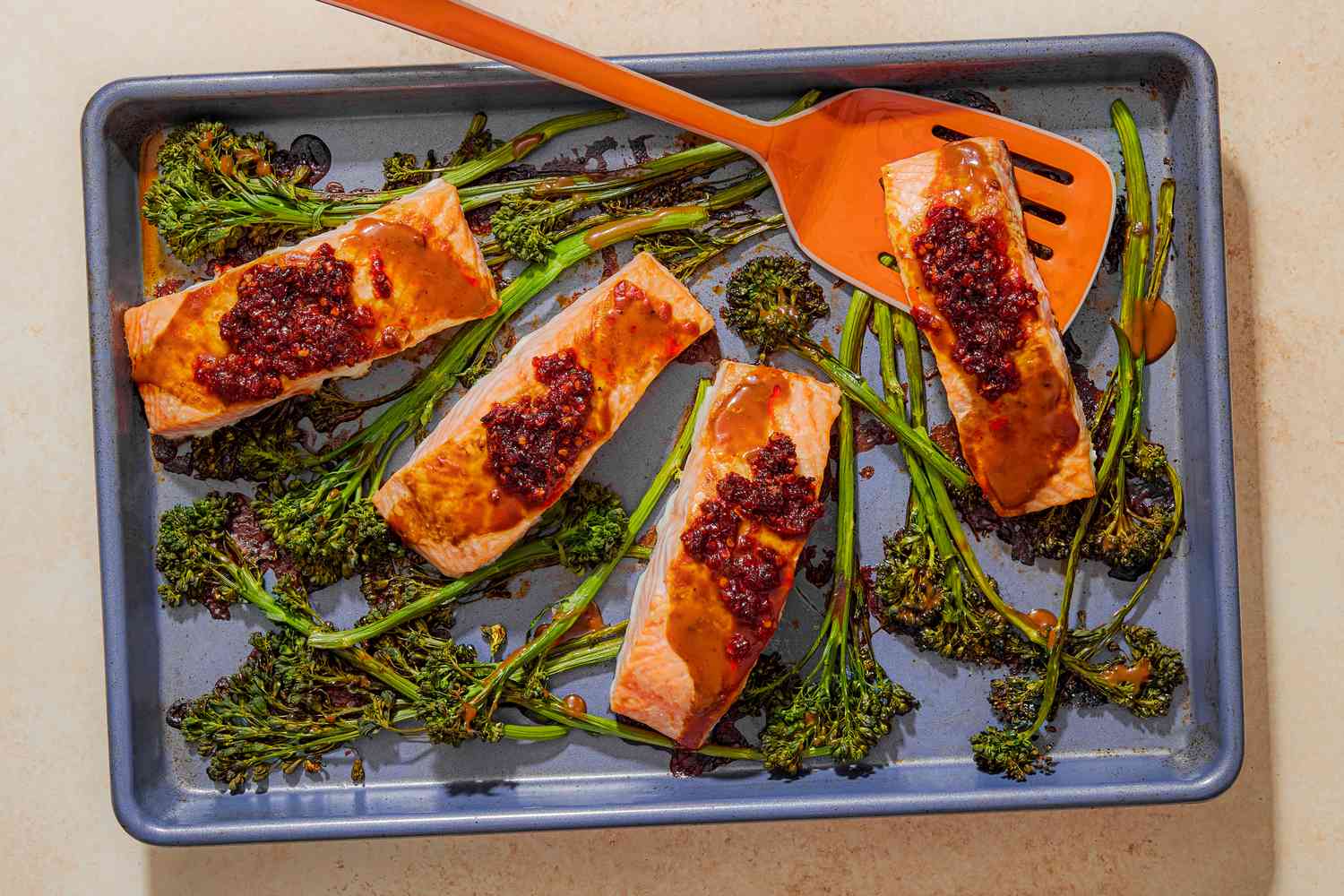 Overhead view of a baking sheet of four cooked salmon filets topped with chili crisp and stems of broccolini with an orange spatula under a piece of salmon