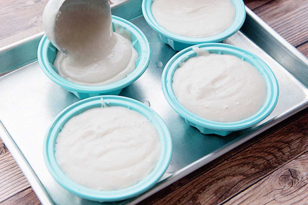 Homemade tembleque is being scooped into four teal molds that are set on a baking sheet. A soup ladle is pouring the pudding into the mold on the top left.