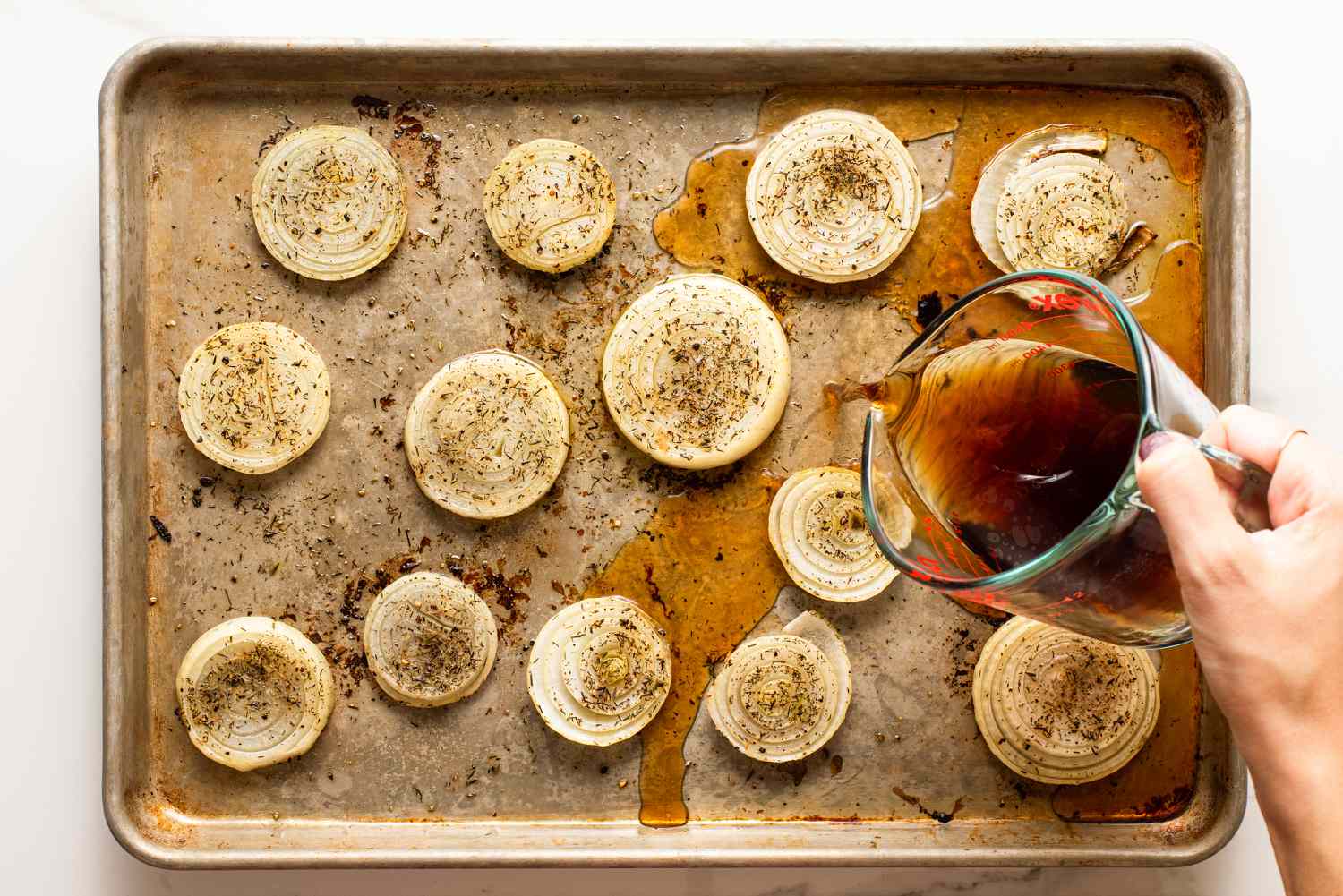 overhead view of raw onions, oiled and seasoned on a sheet-pan, hand pouring broth mixture into the sheet-pan for Melting Onions recipe