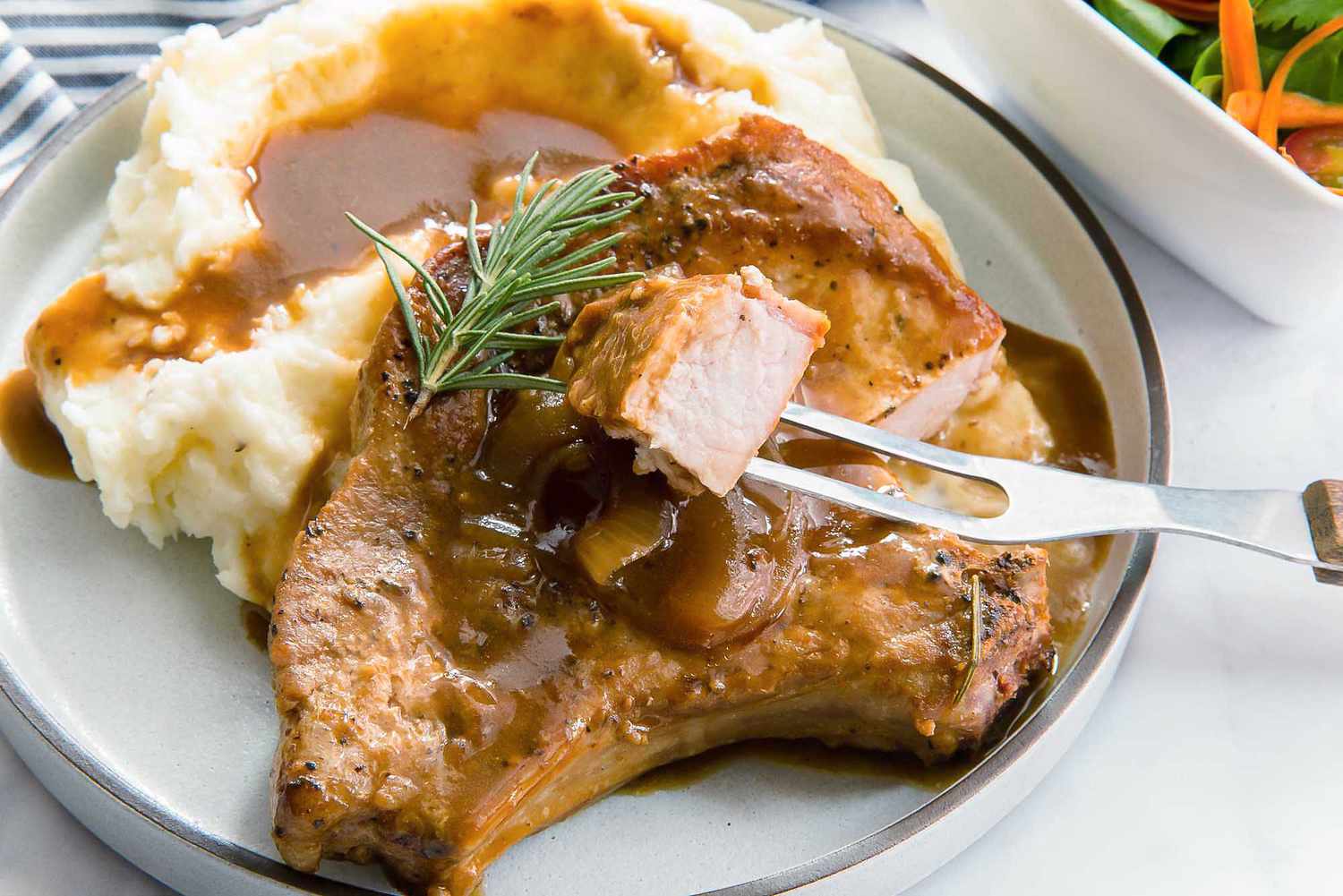 A gluten free smothered pork chop with a pile of mashed potatoes covered in gravy on an ivory plate with a striped linen nearyby and a side salad.