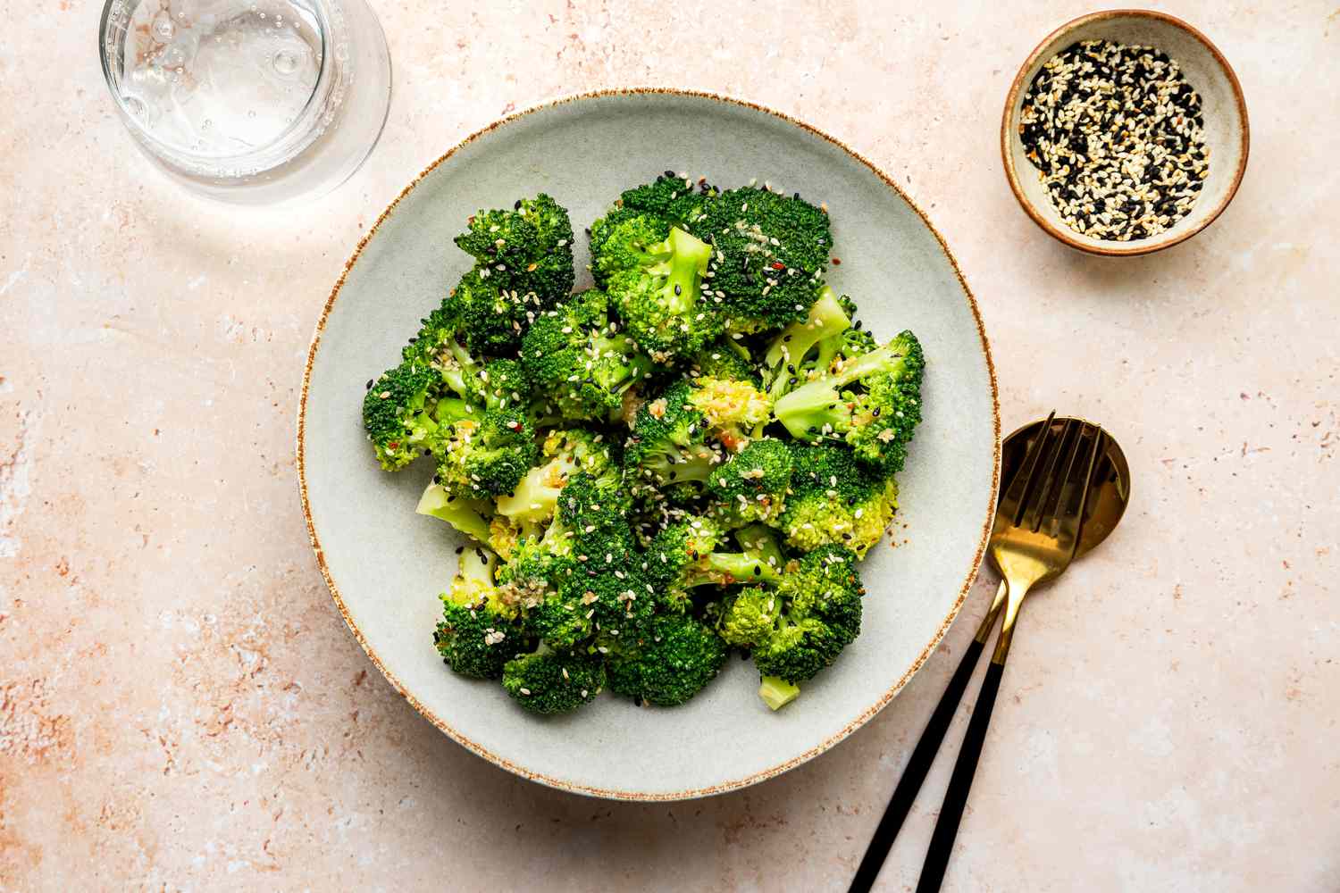 Bowl of Microwave Broccoli with Sesame Dressing Next to Utensils, a Glass of Water, and a Bowl with More Sesame Seeds