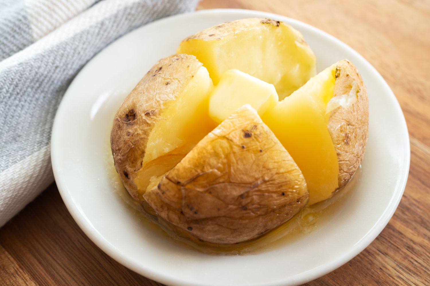 Angled view of a small white plate with a baked potato sliced in quarters and topped with butter