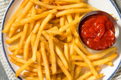 Overhead view of a white plate of french fries with a metal bowl of tomato ketchup