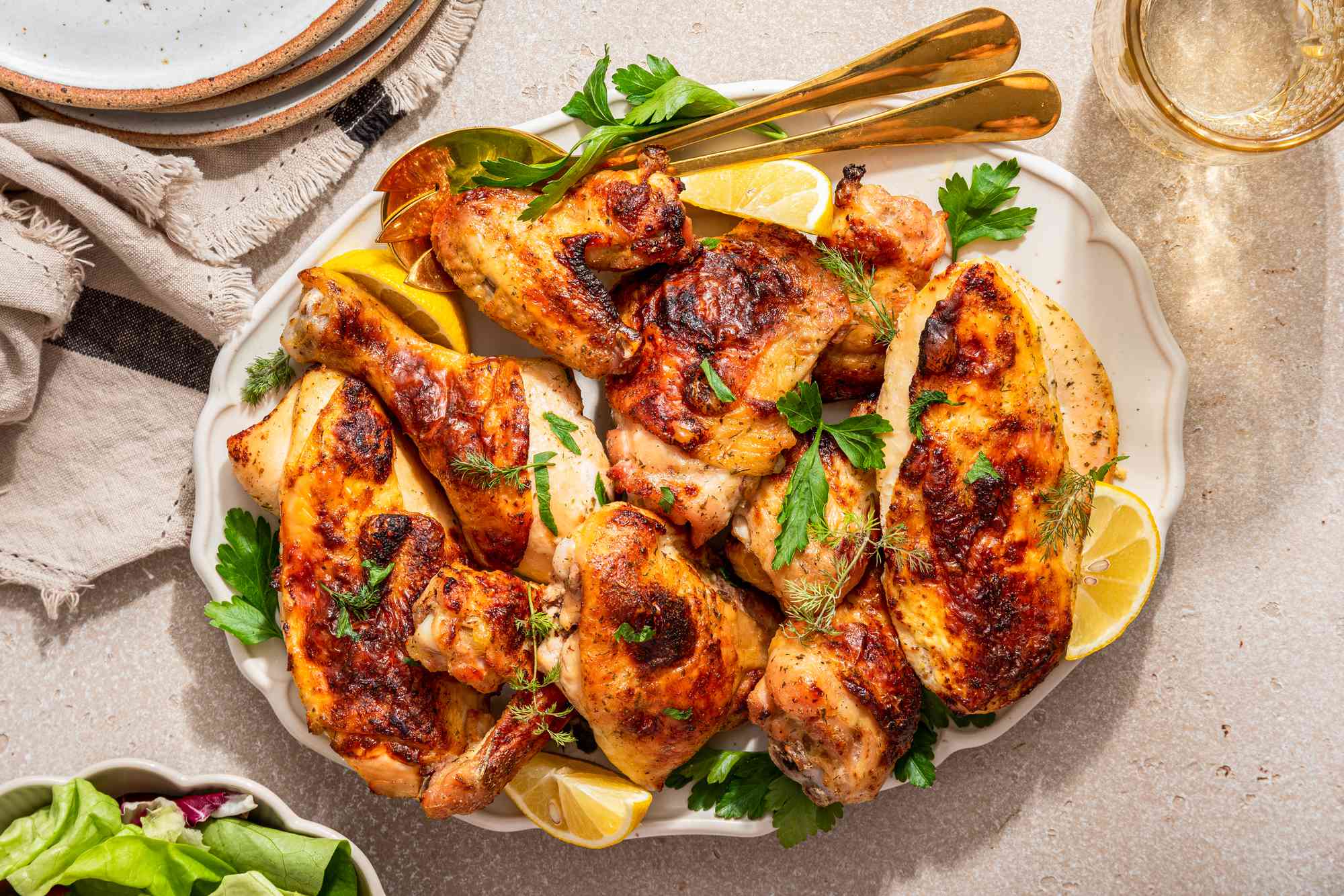 Ranch chicken topped with fresh herbs and lemon wedges on a platter at a table setting with a stack of plates, a bowl of salad, and a glass