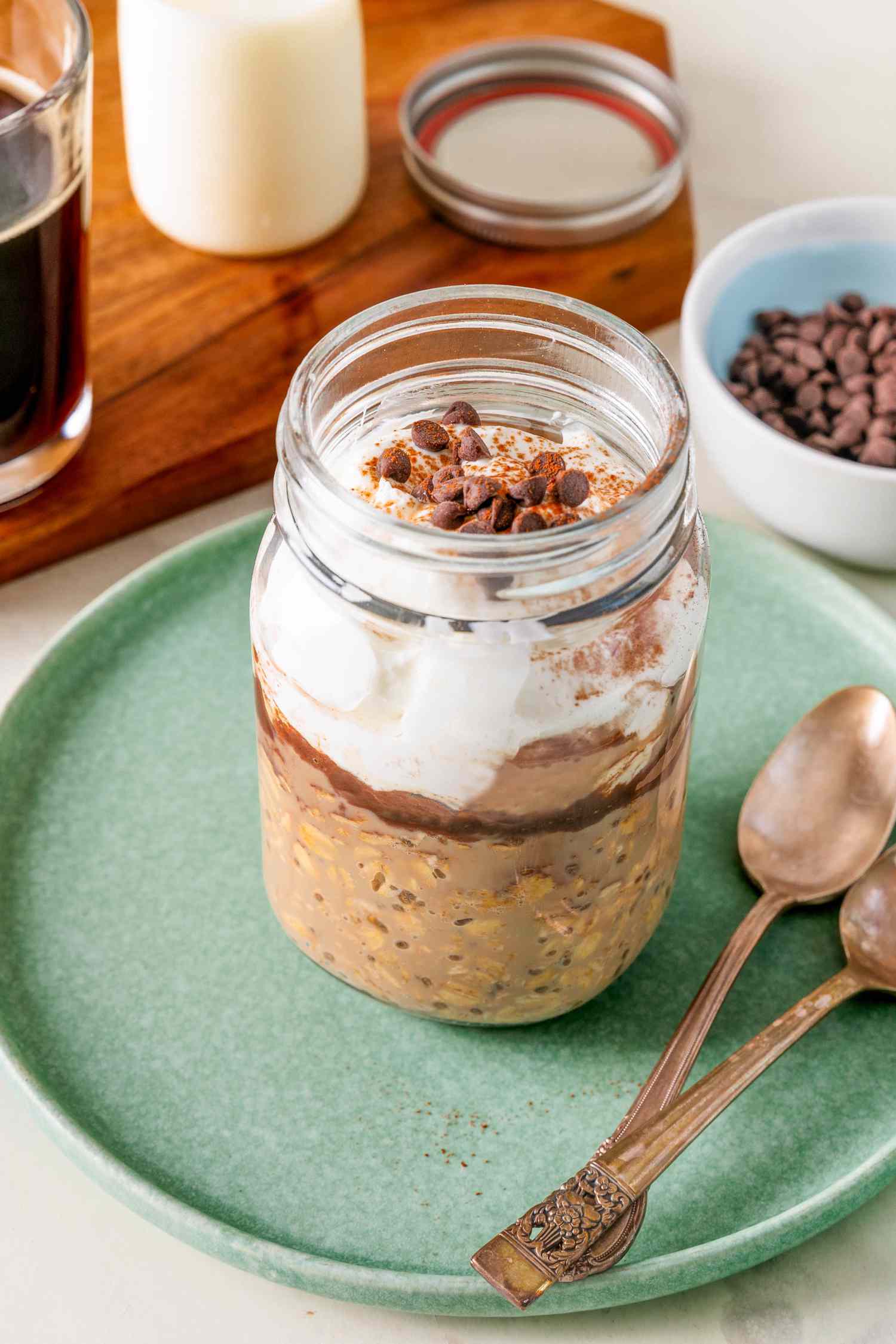 Jar of tiramisu overnight oats topped with Greek yogurt, mini chocolate chips, and cocoa powder on a small plate with two spoons, and in the surroundings, a bowl of mini chocolate chips, and a wooden board with a glass of coffee, a glass of milk, and the jar lid