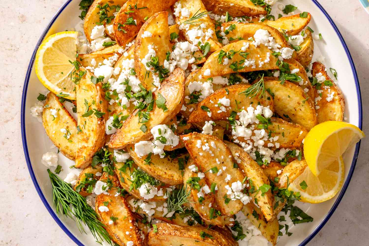 Overhead shot of a serving plate with Greek-style baked potato wedges, garnished with crumbled feta, dill and lemon wedges